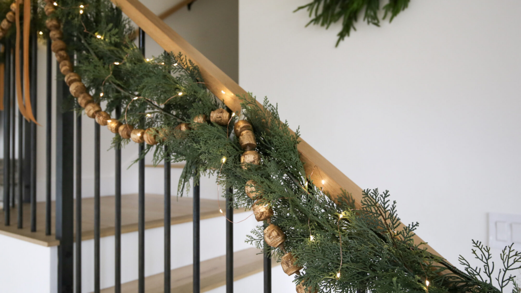 Close-up of a holiday staircase garland with evergreen branches, golden bead accents, and warm twinkling lights on a modern wood and black railing.